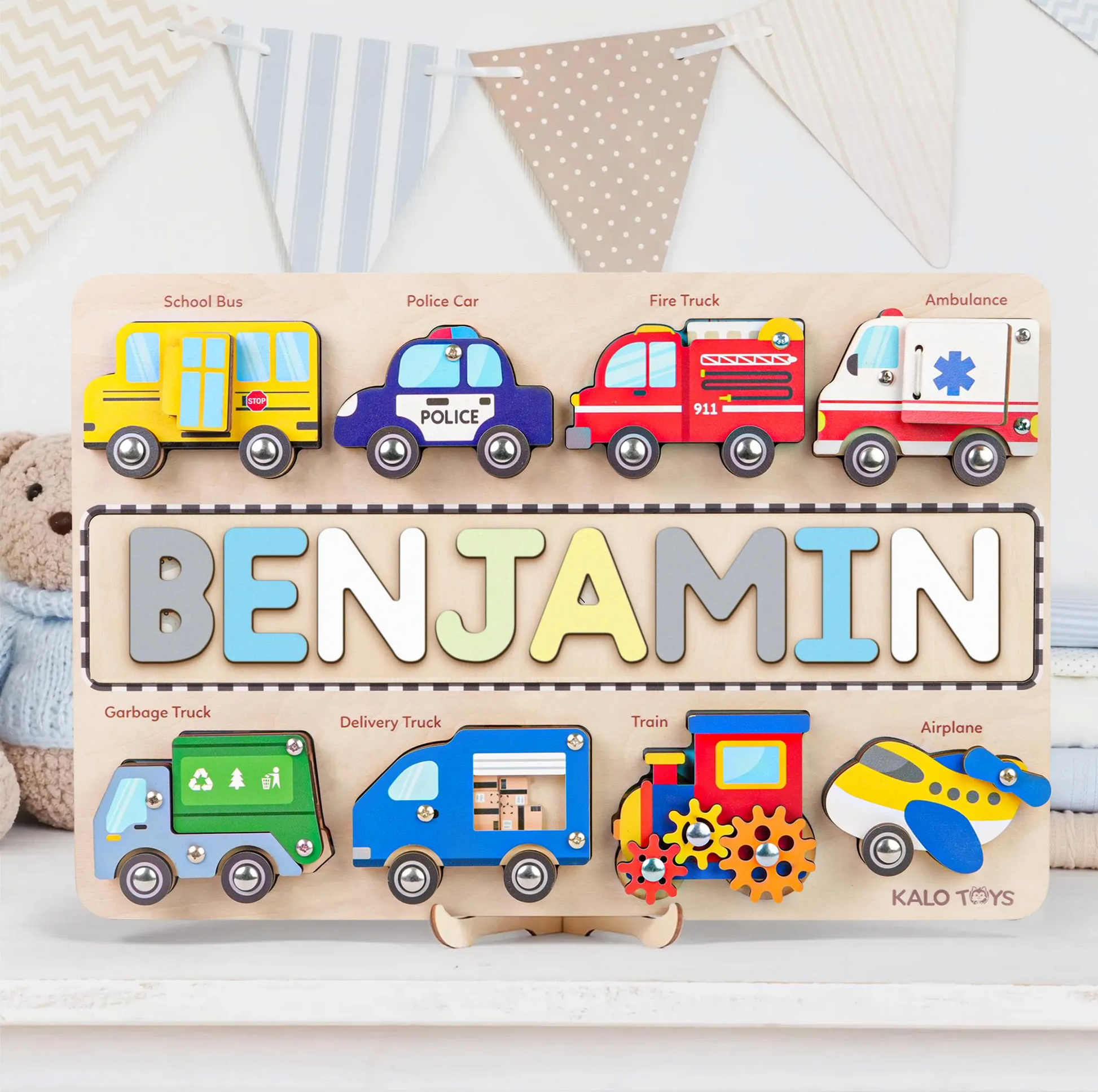 A personalized wooden name puzzle for children, featuring colorful vehicle pieces such as a school bus, police car, fire truck, and ambulance, as well as a garbage truck and delivery truck. The puzzle displays the name 'BENJAMIN' in large wooden letters.
